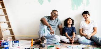 A Black father sits with his Asian partner & their child by a wall they've been painting mint green. They are all smiling and laughing