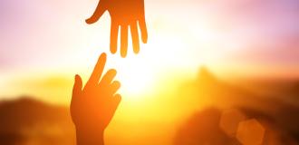 Two silhouetted hands reach toward each other from the top and bottom of the image in front of a sunrise. 