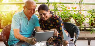 An elderly South Asian couple sits at a table on their balcony looking at their laptop and smiling.