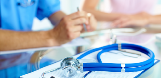 A doctor sits with a patient at a table. On the table there is a stethoscope and a clipboard.