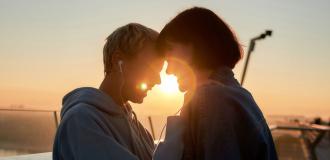 Two young white women stand together closely, laughing. They share headphones and stand in a close embrace