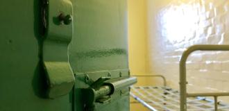 A green jail cell door is partially open, revealing the metal bed frame in the cell.