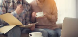 Two old white people sat at a coffee table reviewing some papers over a cup of coffee. A cup of coffee and laptop are in the foreground. 