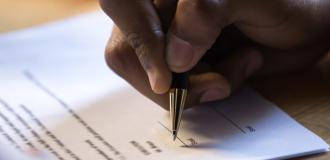 A black male hand is signing a piece of white paper with a gold pen