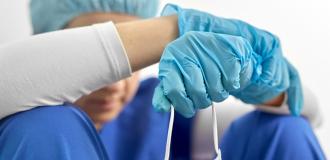 A doctor sits on the floor slumped against the wall. Their arms rest on their knees while their head bows. They're wearing scrubs, blue latex gloves and are holding a mask. 