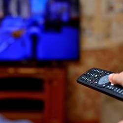 A close-up of a caucasian hand holding a TV remote, pointing it at a television in the background