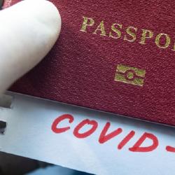 A hand wearing a medical glove holds a burgundy passport. A slip of paper is peeking out of the passport, it reads 'Covid-19' in red writing.