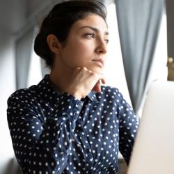 A young woman with black hair wearing a dark blue shirt sits in front of a laptop. She looks off into the distance pensively. 