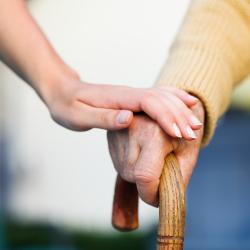 A carer supports the an elderly person's hand. They are holding a cane.