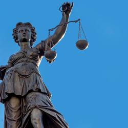 A bronze statue of Lady Justice against a clear blue sky. She is holding a pair of scales in one hand and a sword in the other and is blindfolded.