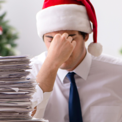 a man wearing a santa hat, holding his head in his hands with a stack of papers on a desk in front of him 