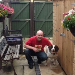 An image of Lee Arnold against a grey background. Lee is a white bald-headed man. He is wearing a red tshirt, crouching next to a dog. He's smiling and seems happy.  