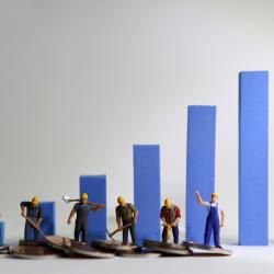 Small models of workers shovelling coins in front of blue bar charts. 