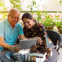 An elderly South Asian couple sits at a table on their balcony looking at their laptop and smiling.