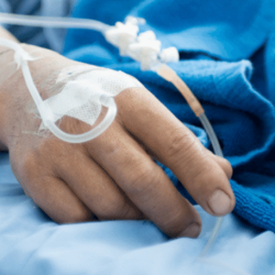 A white person lays in a hospital bed, they are covered by a blue blanket and have an IV drip