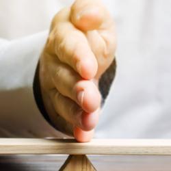 A white hand is on a seesaw between two wooden blocks. The one on the left is green, the one on the right is red. The seesaw is in balance.