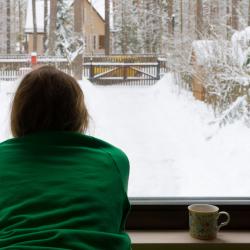 A person with brown hair and an emerald green jumper can be seen from behind, looking out of a large window onto a snowy back garden. There is a cup of tea next to them. 