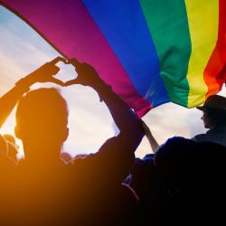 A silhouette of a person making a heart with their hands under a large rainbow flag that's held by someone in the foreground.