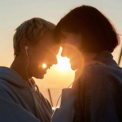 Two young white women stand together closely, laughing. They share headphones and stand in a close embrace