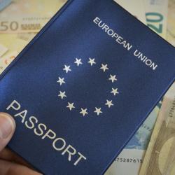 A blue EU passport being held in front of Euro banknotes.