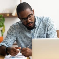 A Black man in a blue shirt sits in front of a laptop and looks at it seriously while making notes on a piece of paper.