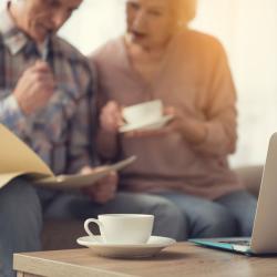 Two old white people sat at a coffee table reviewing some papers over a cup of coffee. A cup of coffee and laptop are in the foreground. 