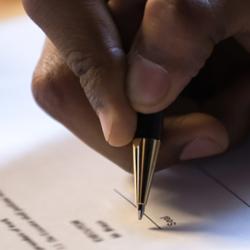 A black male hand is signing a piece of white paper with a gold pen