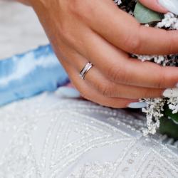 A close up of a white woman's hand resting on top of a wedding bouquet made of light purple and white flowers. The hand is wearing a silver wedding band.