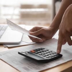 A stack of papers with a calculator on it are on a coffee table. A hand is holding a bit of paper and another hand is pressing a button on the calculator