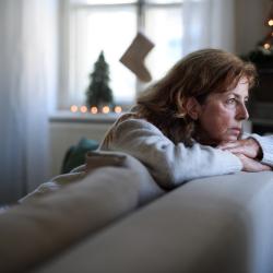 A white woman in her fifties sits on a sofa and leans over the back of it, looking sadly through the window. There are a Christmas decorations on the cabinet behind her. 