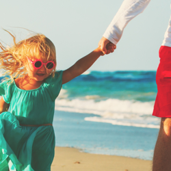 A father holds his daughters hand as they stroll towards the camera along the beach. 