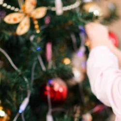 A female white child with blonde hair in pigtails reaches towards a Christmas tree to place a decoration on the branch.