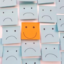 An orange post-it note with a happy face on it amongst many blue post-it notes with sad faces drawn on them