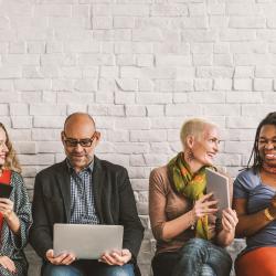 a diverse group of employees sat together on a bench looking at phones, laptops, books