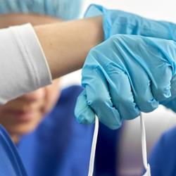 A doctor sits on the floor slumped against the wall. Their arms rest on their knees while their head bows. They're wearing scrubs, blue latex gloves and are holding a mask. 